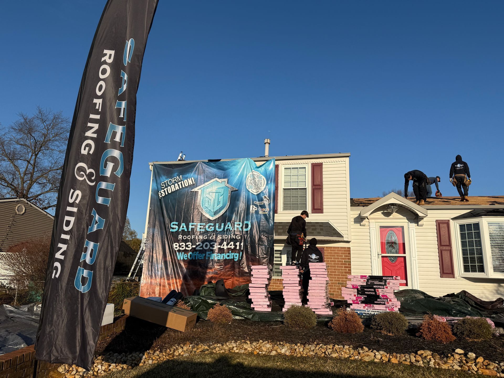 Roofers on a house with pink insulation and a company banner under a blue sky.