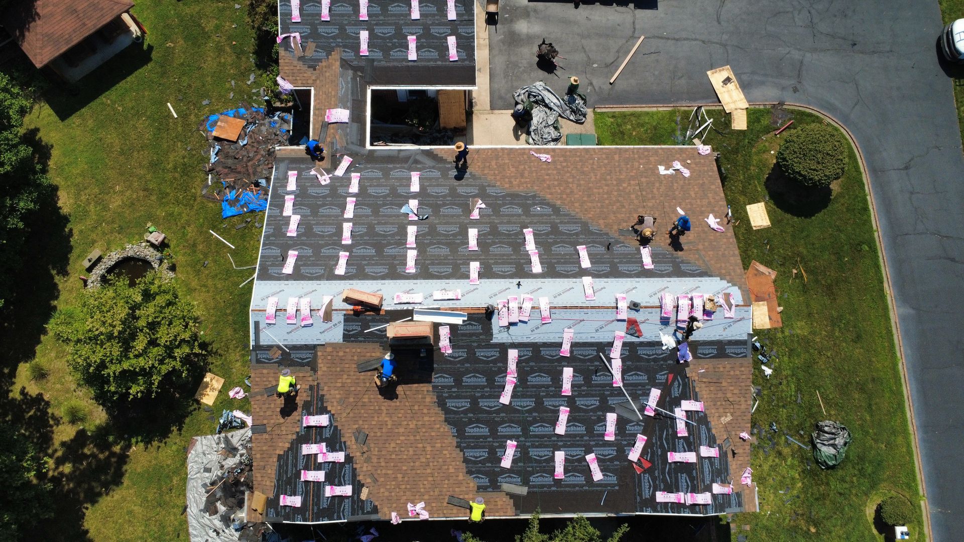 Aerial view of a roof being shingled by several workers. Brown shingles and pink insulation visible.