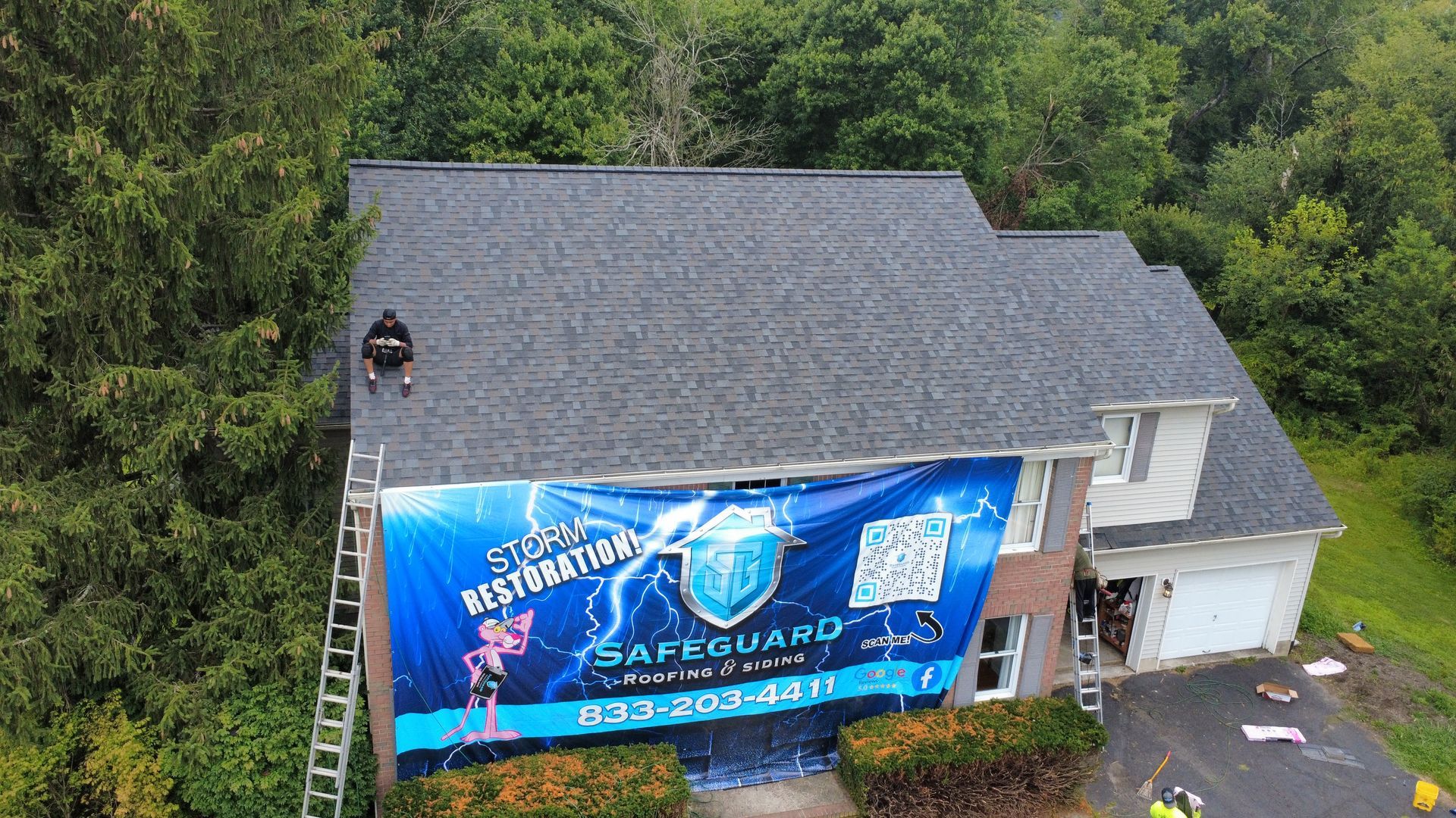 A worker on a house roof with a large banner. Dark gray roof and brick facade. Green trees surround.