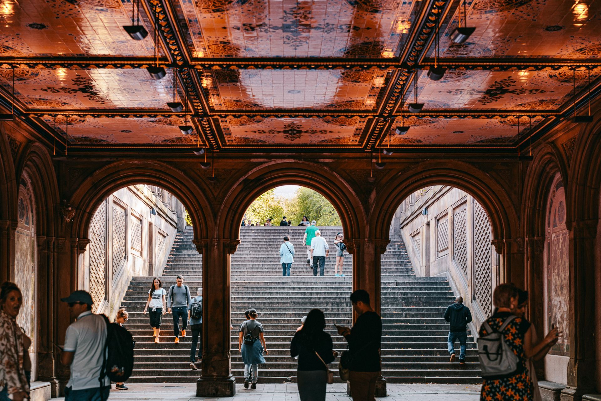 People walking through an arched walkway with ornate ceilings and golden light at Bethesda Terrace