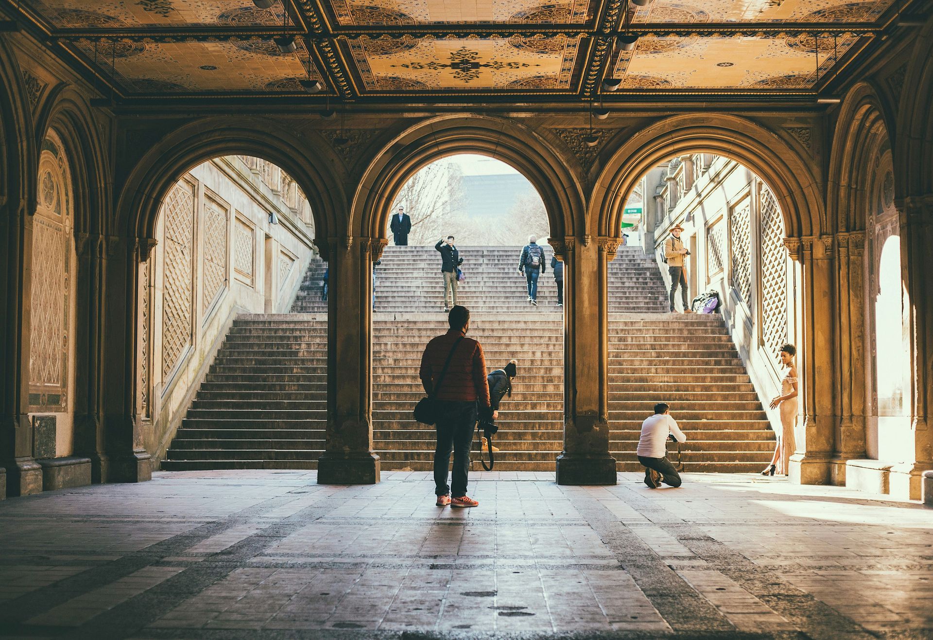 Personas caminando por un pasillo arqueado con techos ornamentados y luz dorada en Bethesda Terrace