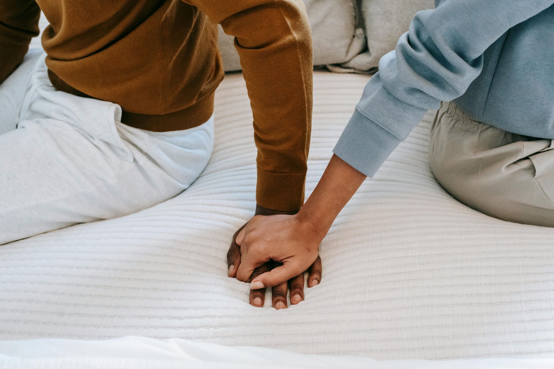 Two individuals on a white bed, hands clasped in a subtle show of mutual support and comfort.