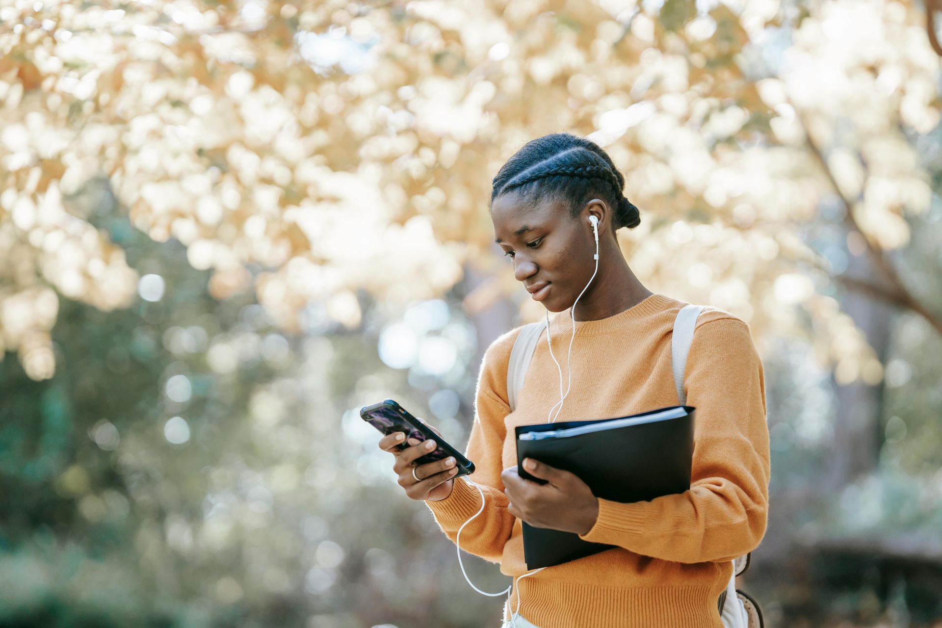 Person wearing earbuds and orange sweater, holding folder and phone while walking in a park.