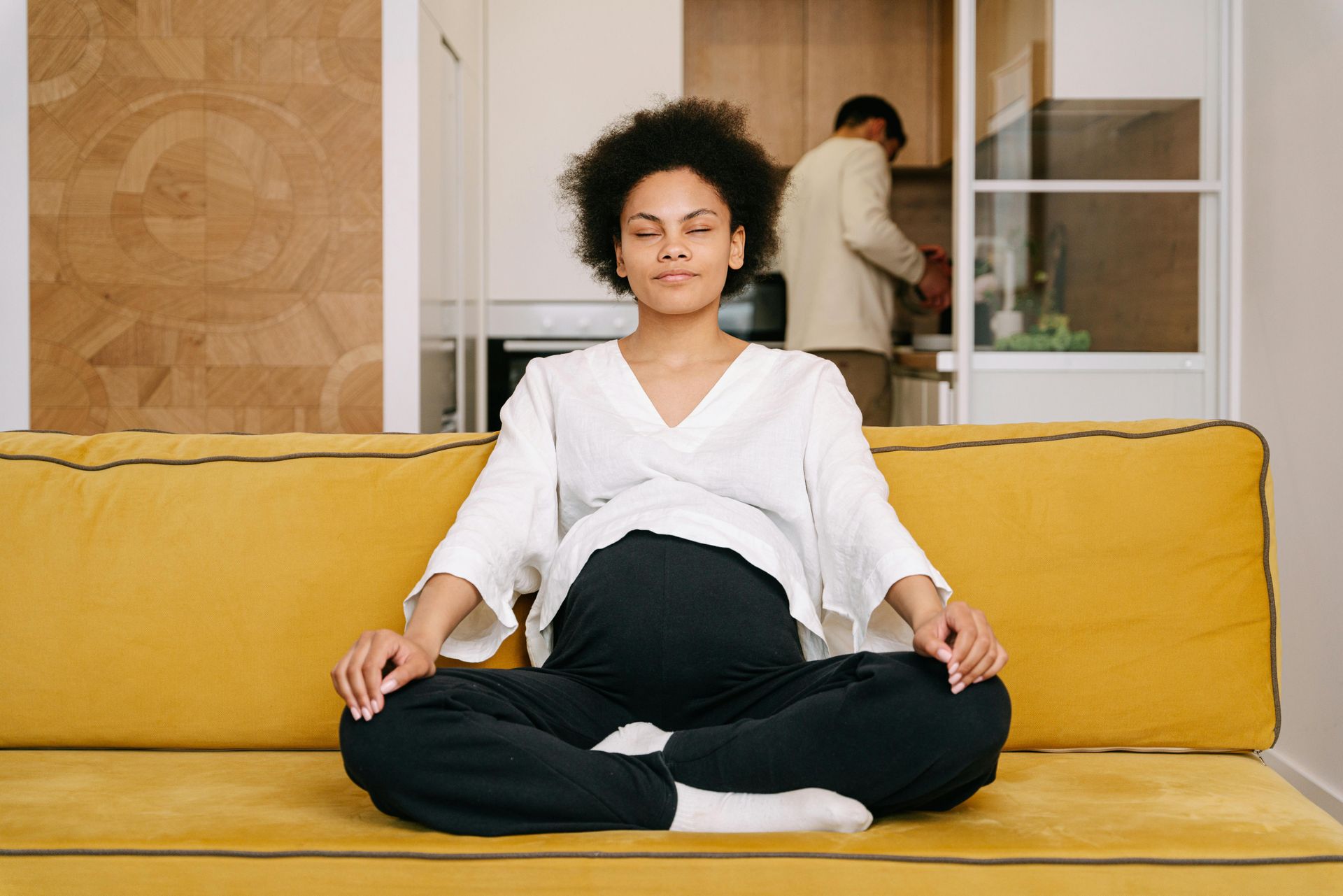 Woman meditating calmly on a yellow couch, eyes closed in a serene living room
