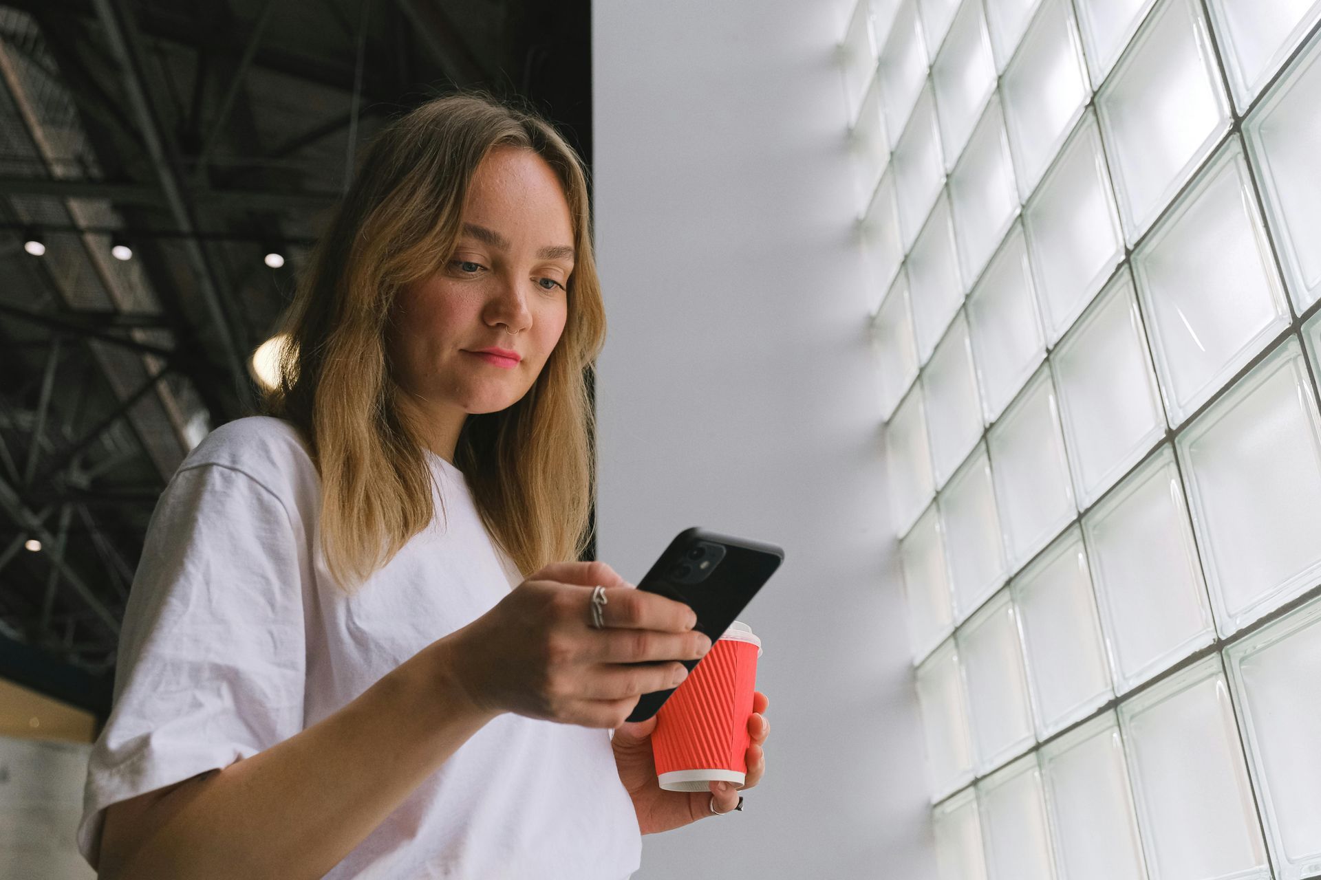 Woman in a white shirt holding a coffee cup and checking her phone by a glass block window.