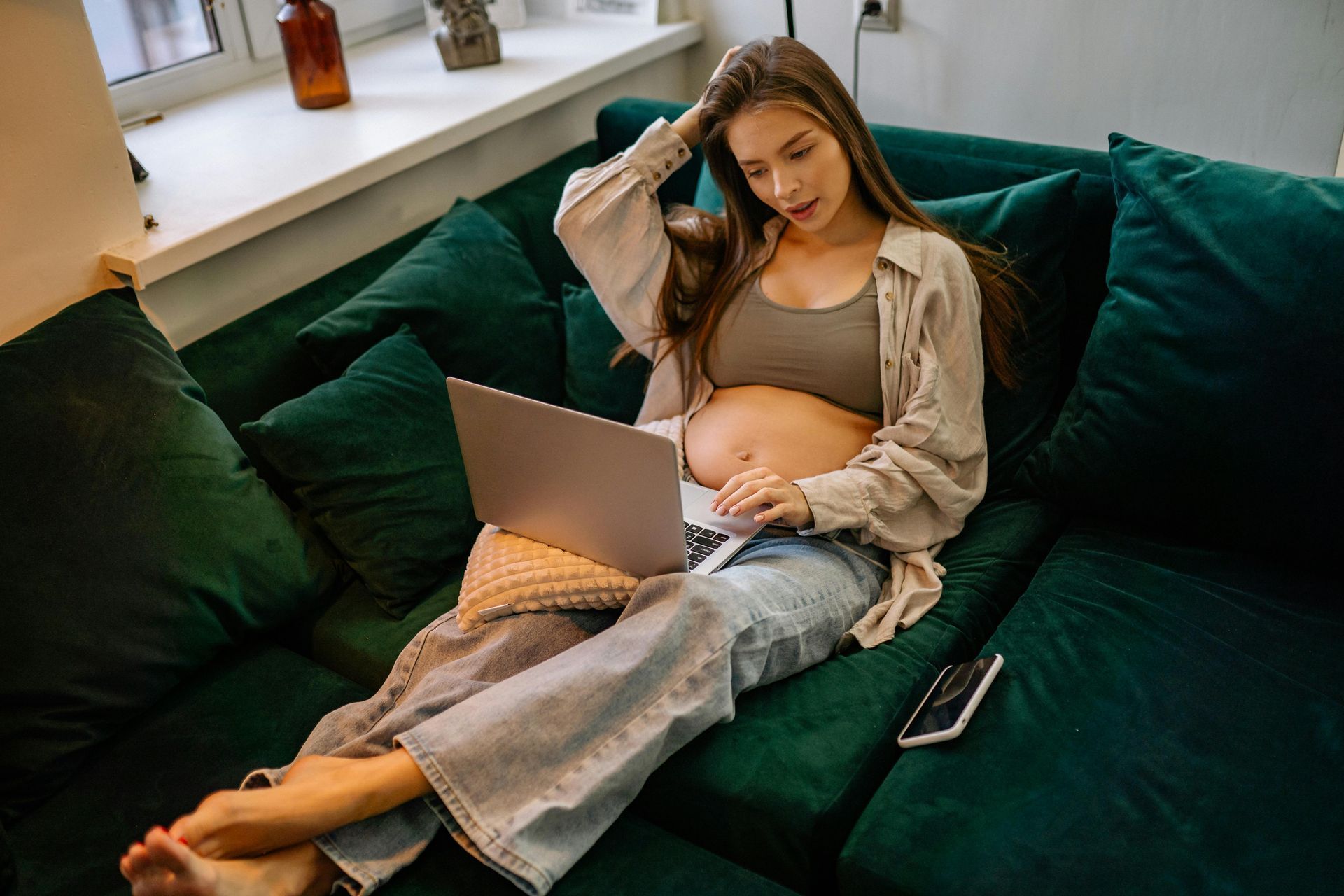 Pregnant woman researching on laptop on a green sofa in our NYC apartment