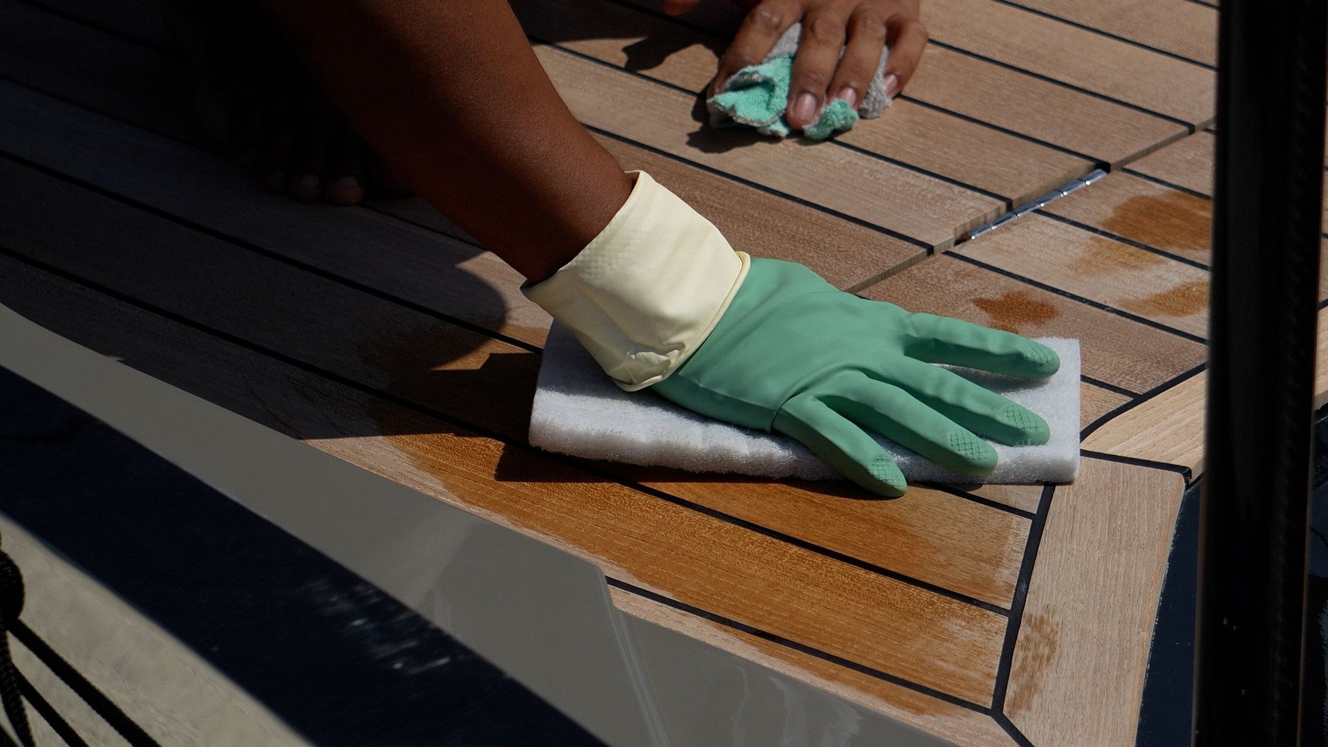 Close-up of crew cleaning and spot-treating a teak yacht deck by hand with a soft pad and cloth, demonstrating careful maintenance techniques to protect the wood surface and seams.