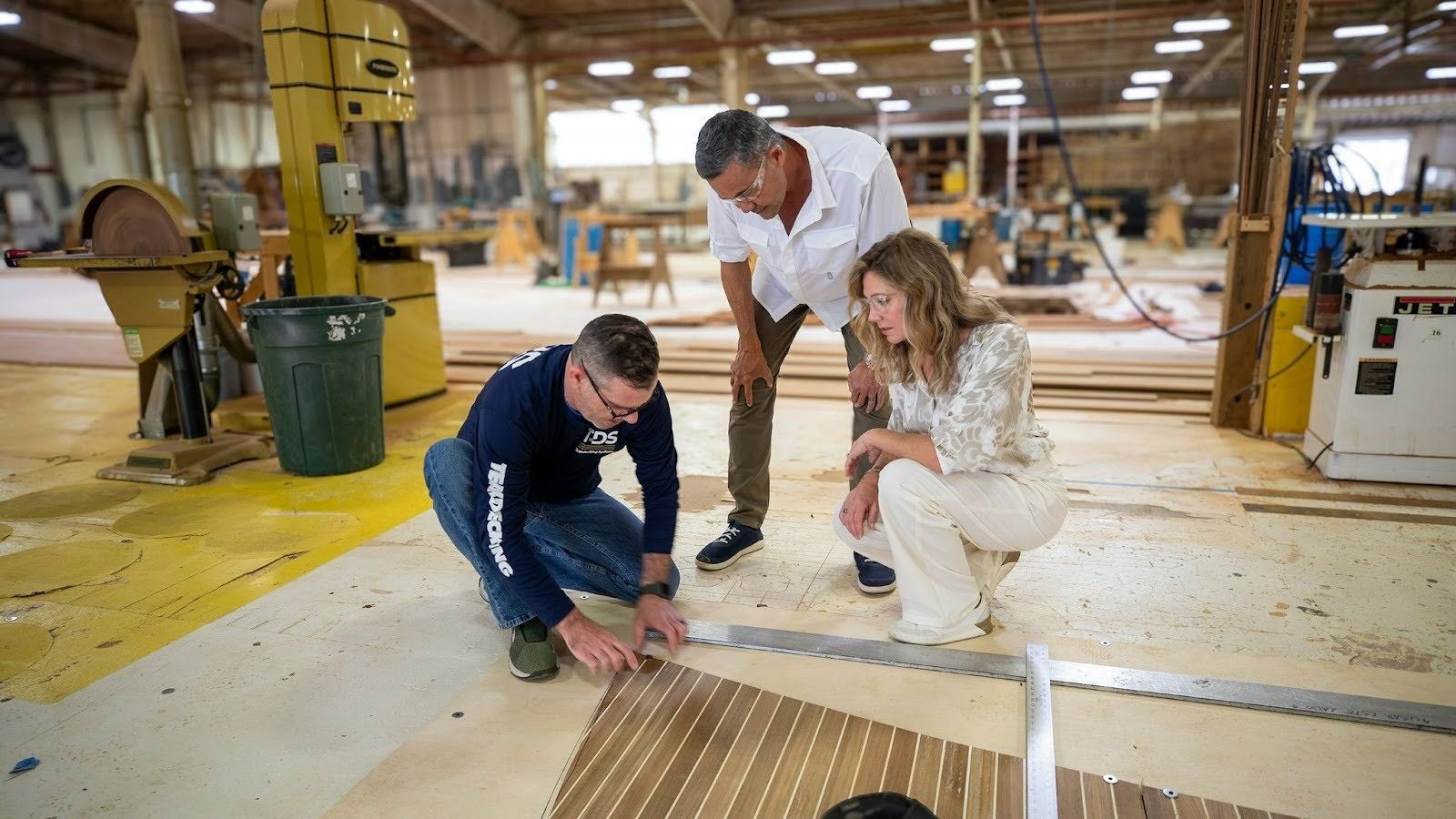 Three people in a workshop reviewing and measuring a decking panel on the floor, with one technician marking measurements while two others observe.