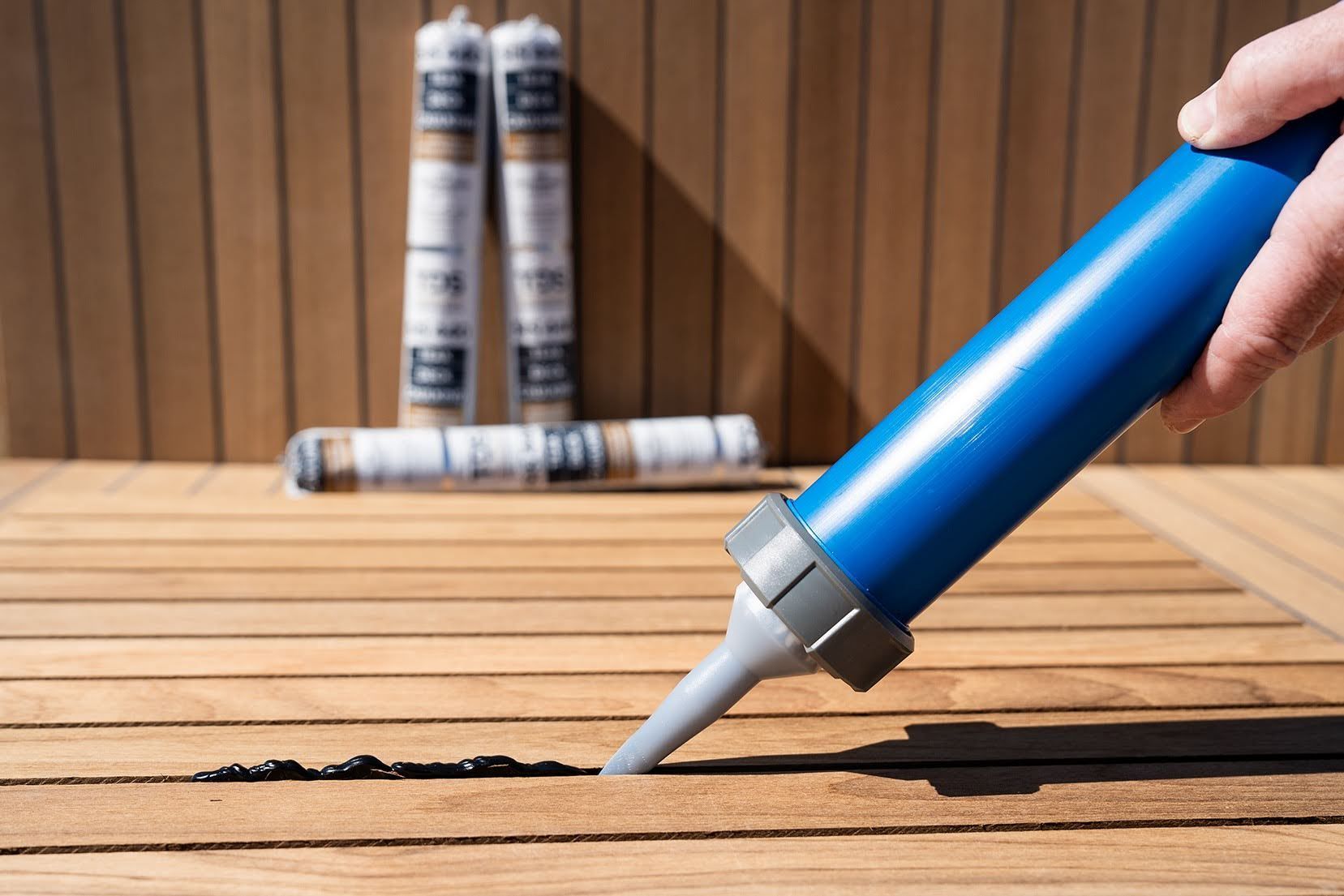 Hand applying sealant from a blue caulking tube into the gap between wooden deck boards, with additional sealant tubes blurred in the background.