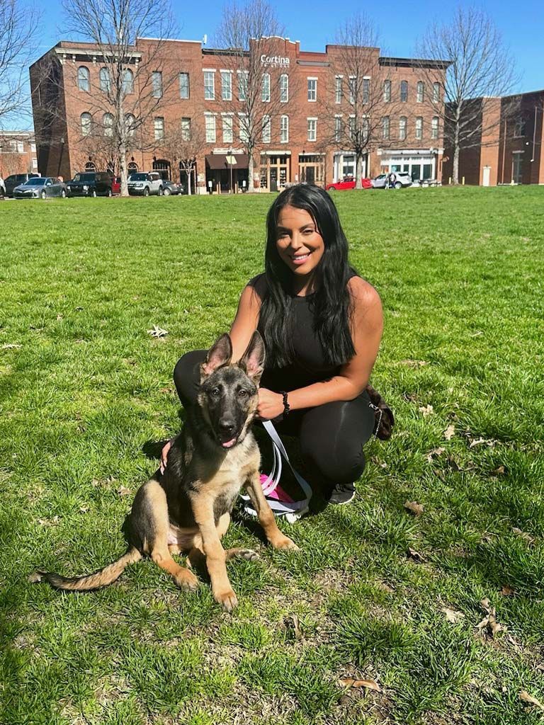 Woman and German Shepherd dog sit on grass in front of brick building. Both are looking at camera.