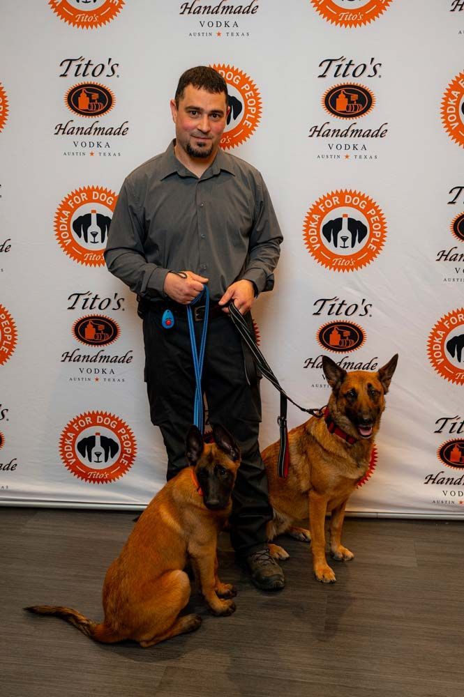 Man with two Belgian Malinois dogs in front of a Tito's Vodka backdrop.