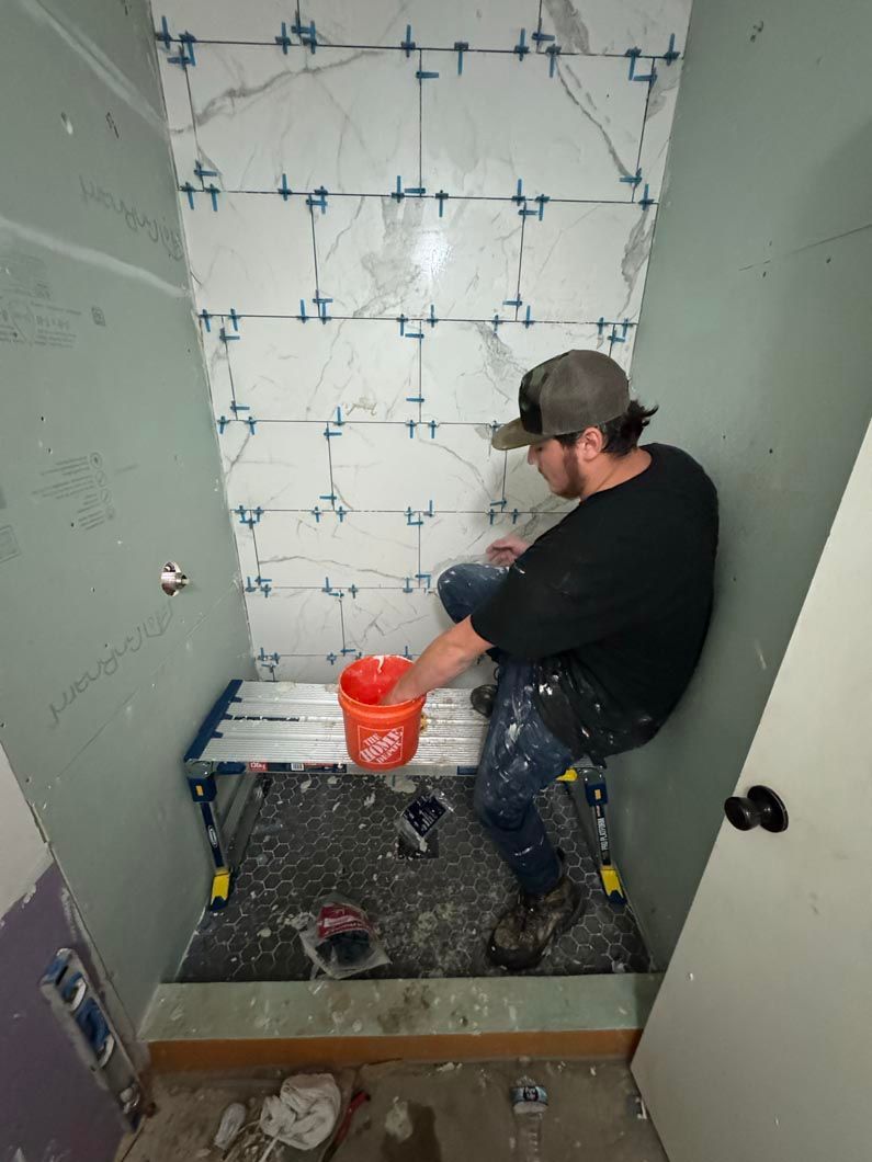 A man is sitting on the floor in a bathroom tiling a wall.