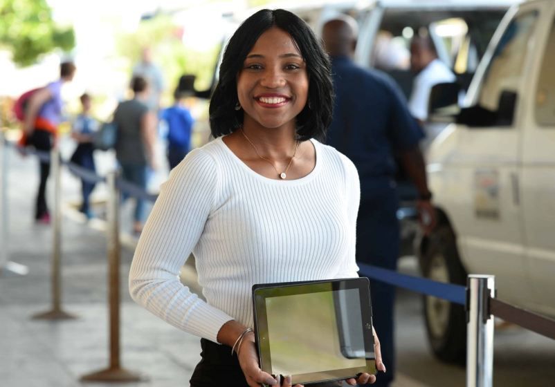 Woman smiling, holding a tablet, standing outdoors near a vehicle and crowd of people.