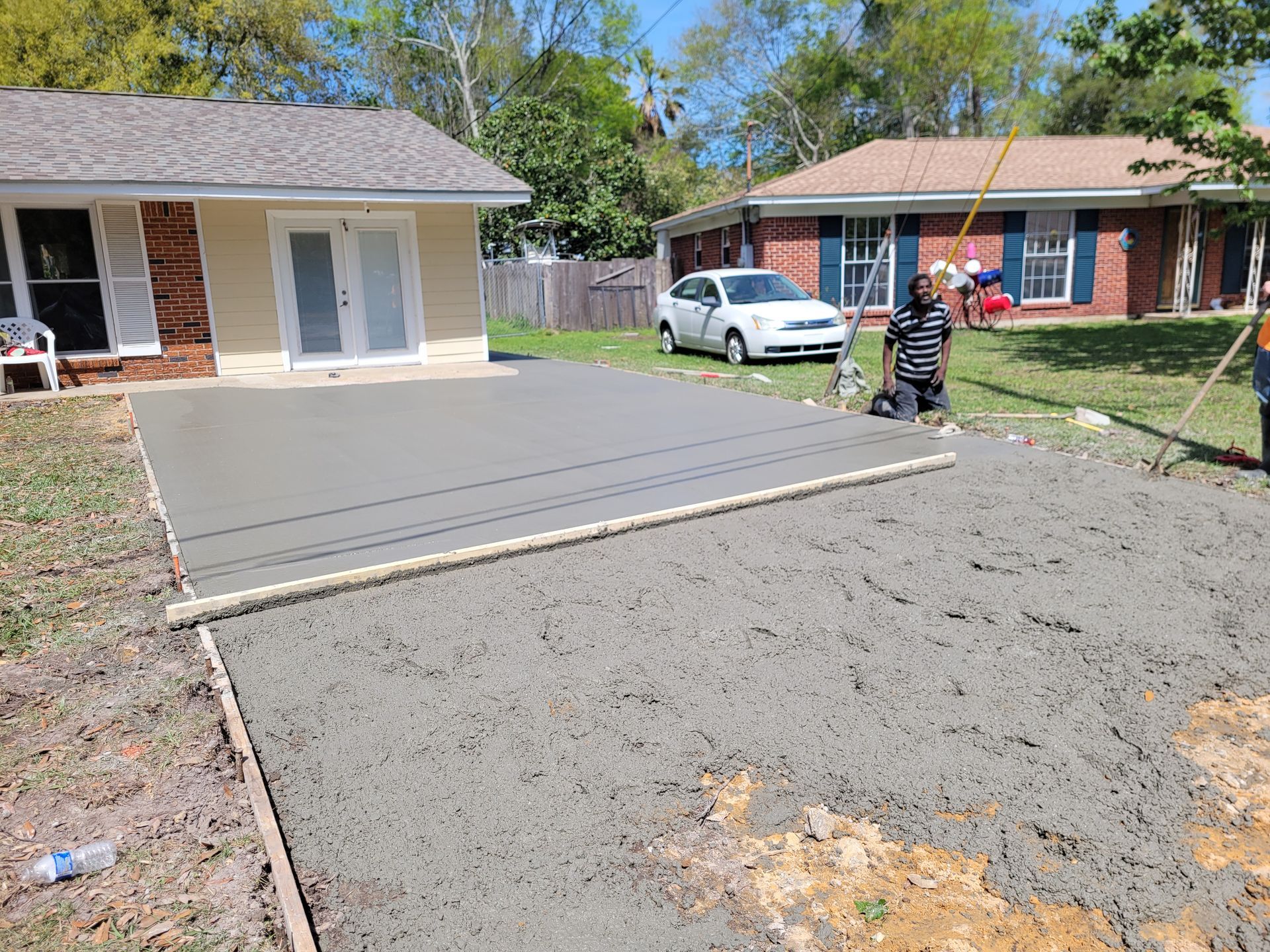A worker smooths a freshly poured concrete patio in front of a residential house on a sunny day.