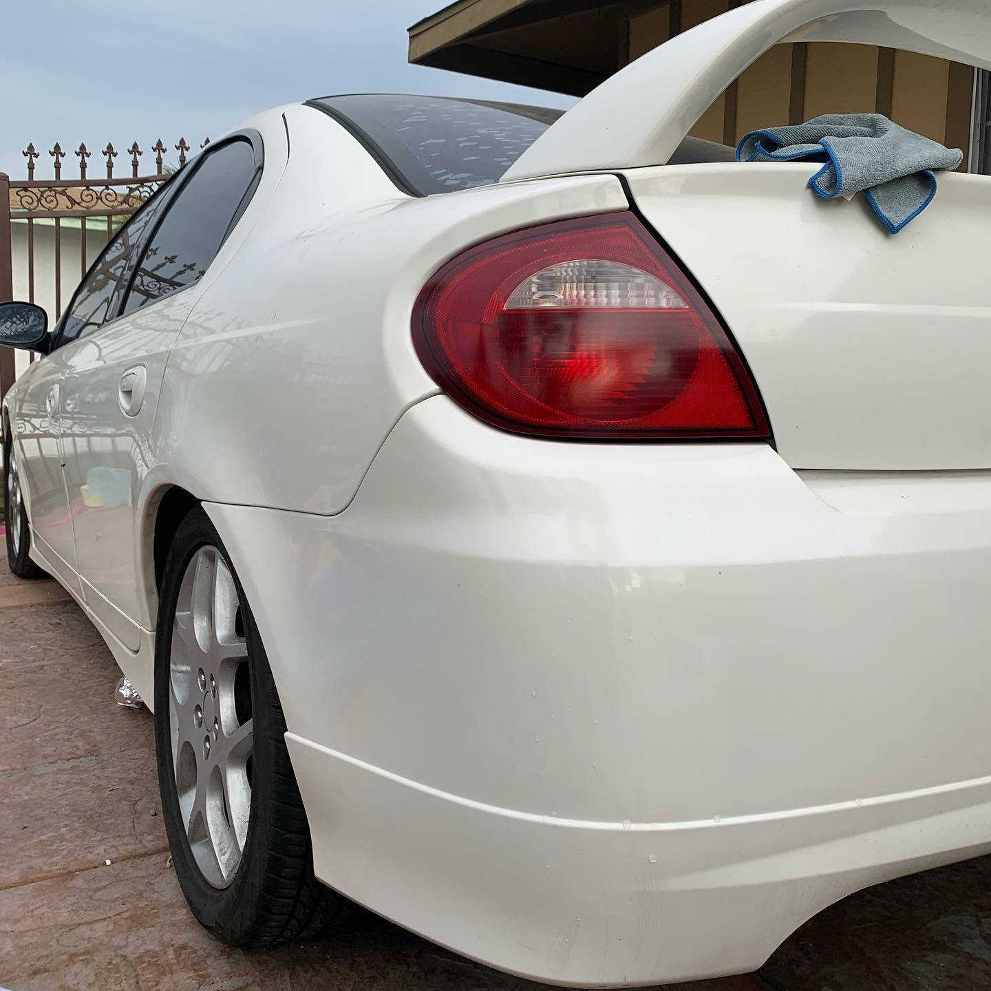 A white car with a red tail light is parked in front of a fence