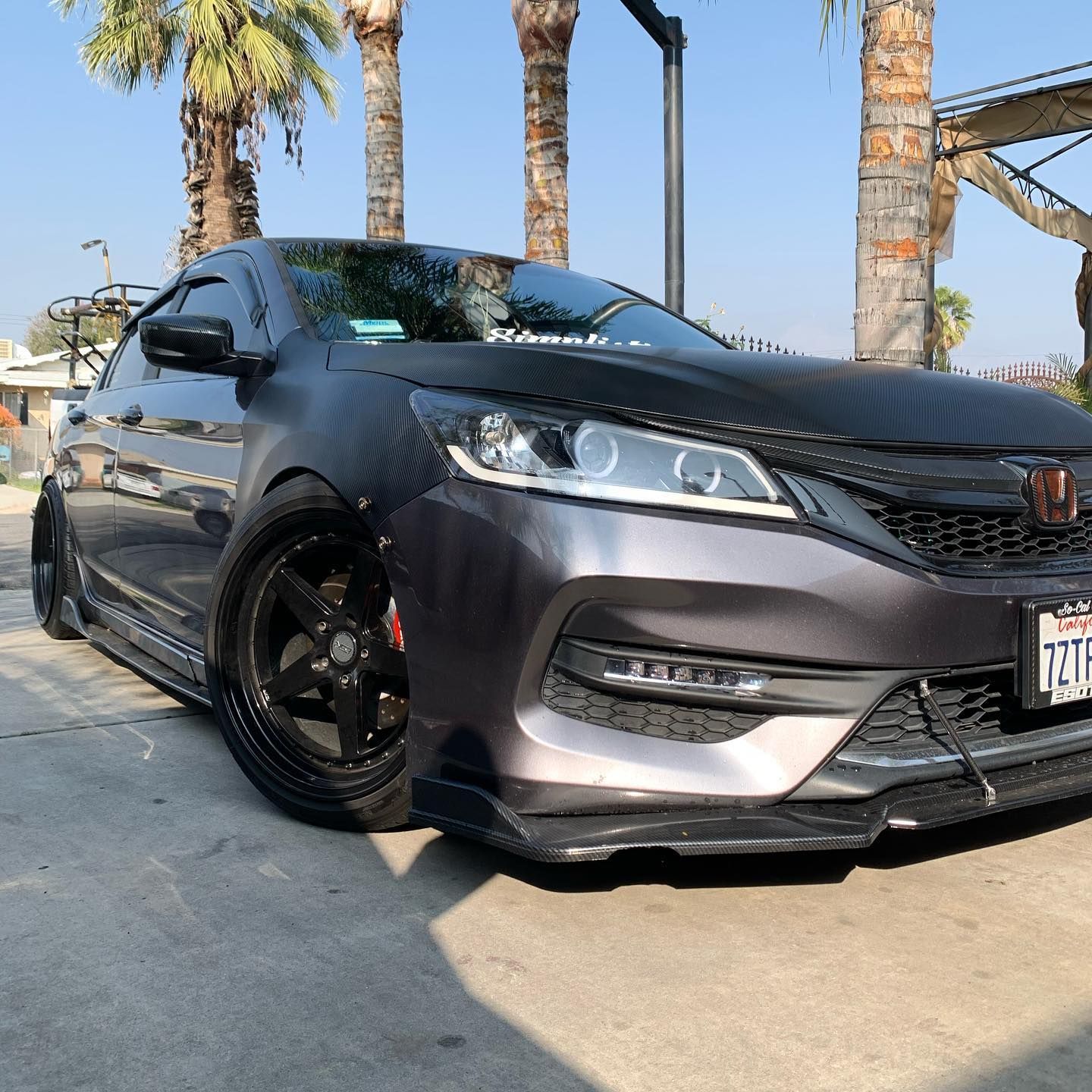 A black car is parked in a parking lot with palm trees in the background.