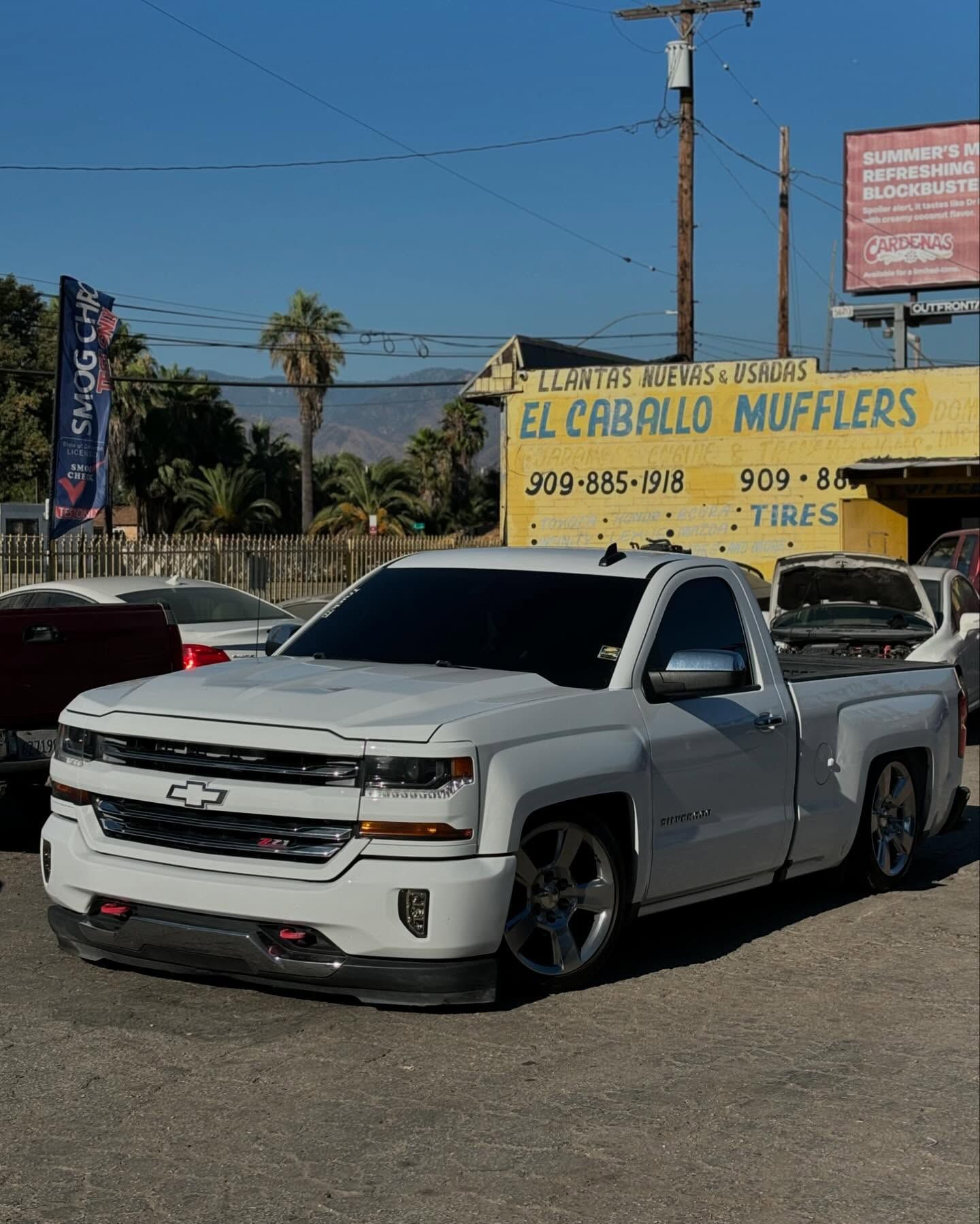 A white truck is parked in front of a yellow sign that says el caballo mufflers
