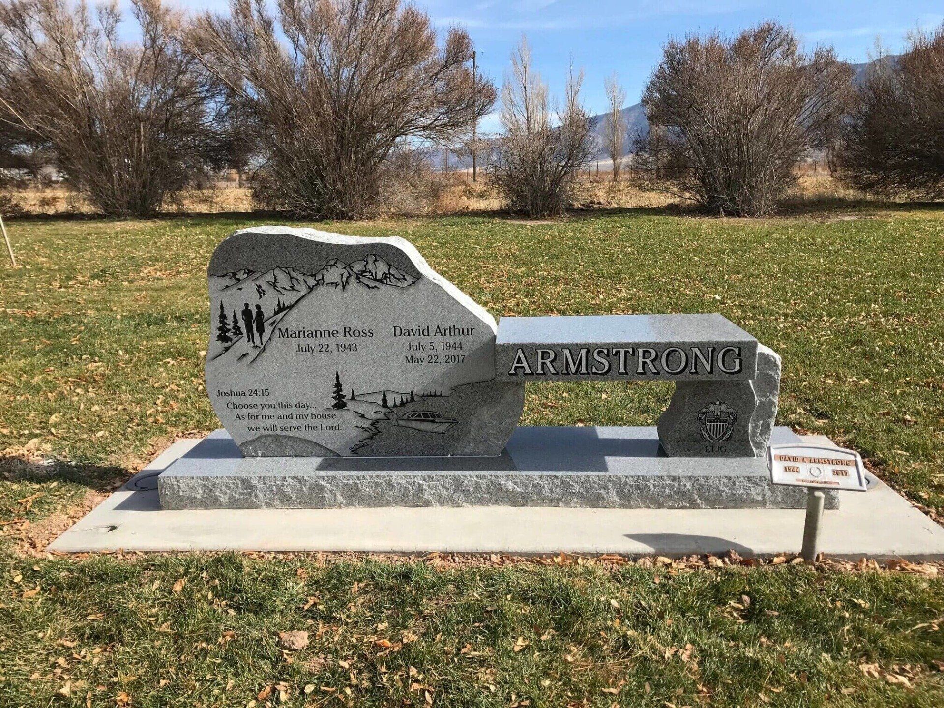 Monument bench engraved with names, dates, and a mountain scene, set in a grassy field under a blue sky.