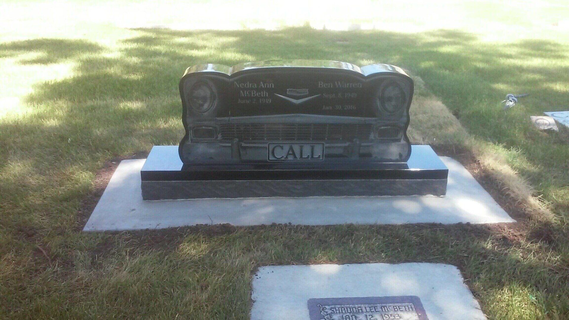 Black headstone in a grassy cemetery, shaped like a classic car, inscribed with the name 