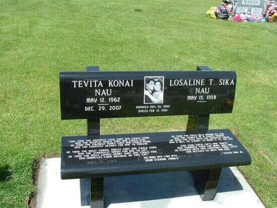 Black memorial bench with names and dates, a photo, and inscriptions on a grassy field.