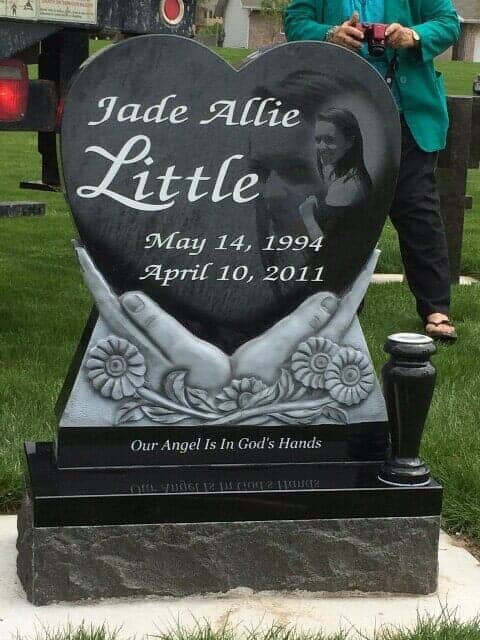 Black heart-shaped gravestone for Jade Allie Little, with hands holding it; a woman stands nearby.