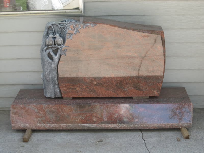 A red granite headstone with carved birds and a base, resting on wooden supports.