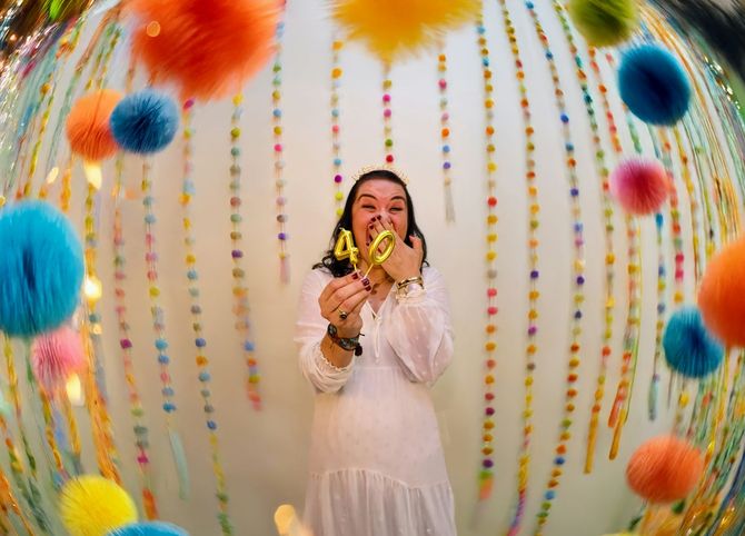 Woman in white dress holding yellow balloons amid colorful hanging pom-poms and streamers