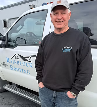 Smiling man leaning against a RainBlock Roofing truck, wearing a black sweatshirt and cap.
