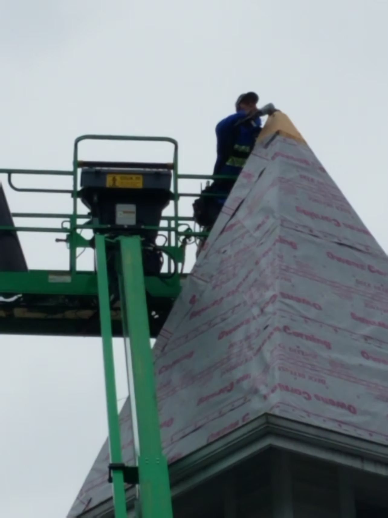 Worker on a green lift installing wrap on a tall pointed structure.