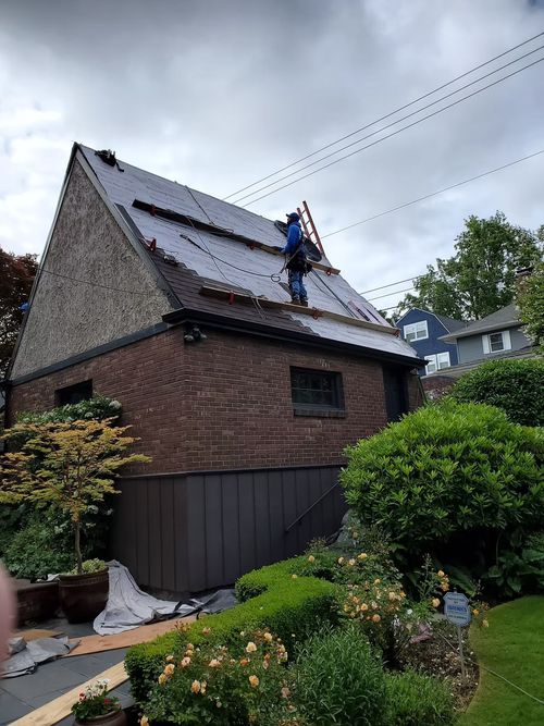 Brick house with steep roof being repaired, worker on the roof, and a garden in the foreground