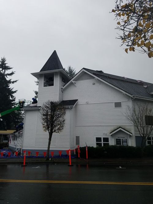 White church with a steeple on a rainy street, with orange cones and a tree in front.