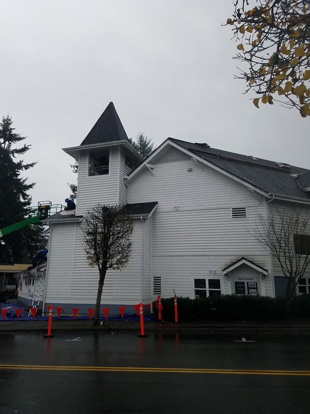 White church with a steeple on a rainy street, with orange cones and a tree in front.