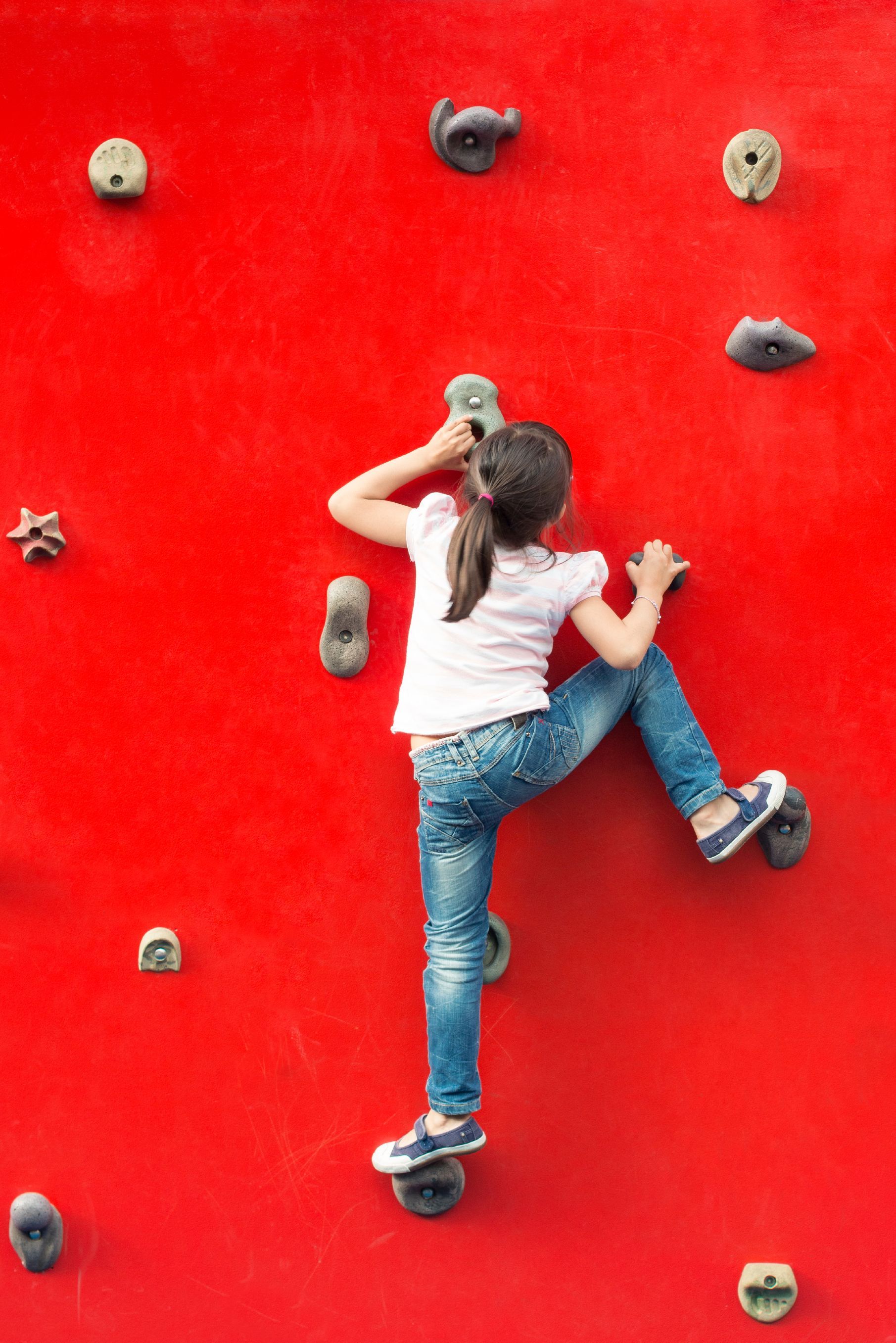 A little girl is climbing up a red climbing wall.