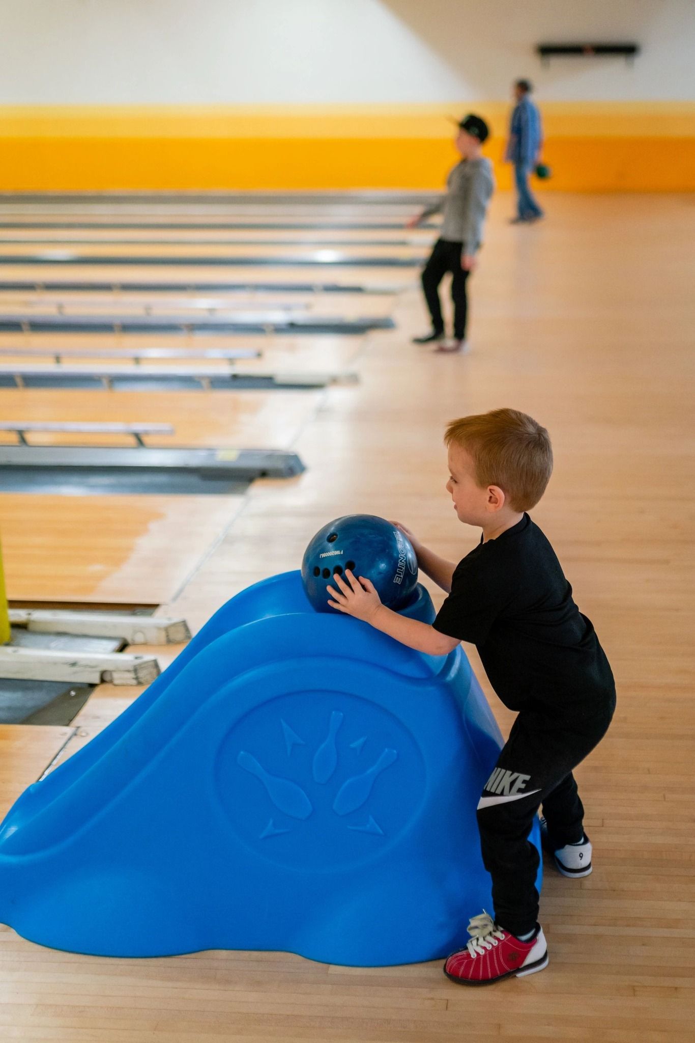 Boy with red hair prepares to bowl using a blue ramp at a bowling alley.