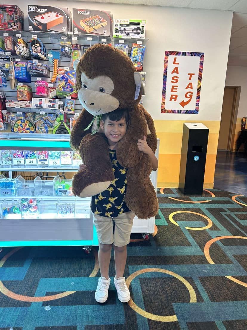 Boy holding a large stuffed monkey in an arcade, standing near a laser tag sign.