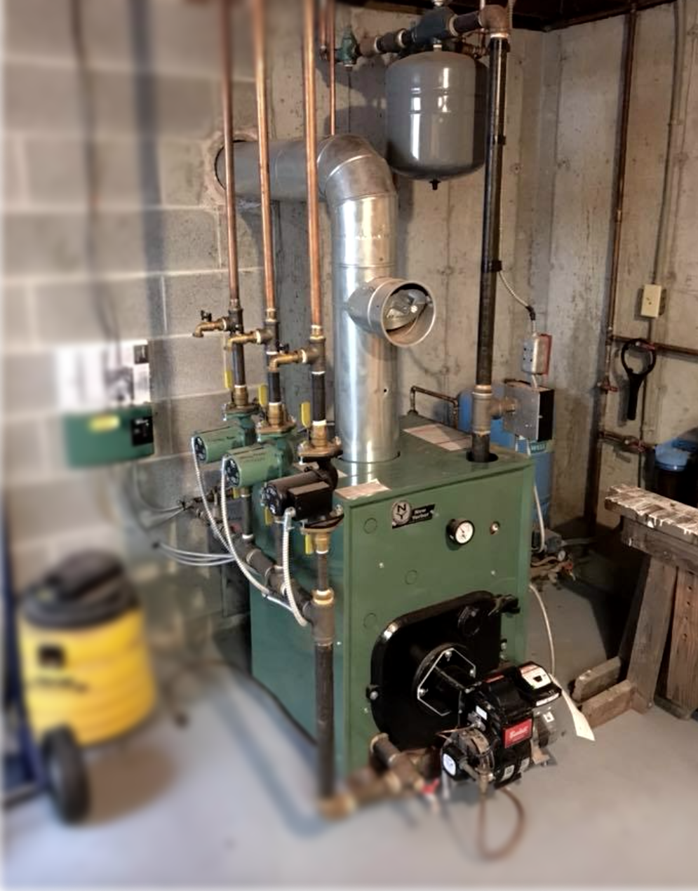 A green boiler is sitting in a basement next to a yellow vacuum cleaner