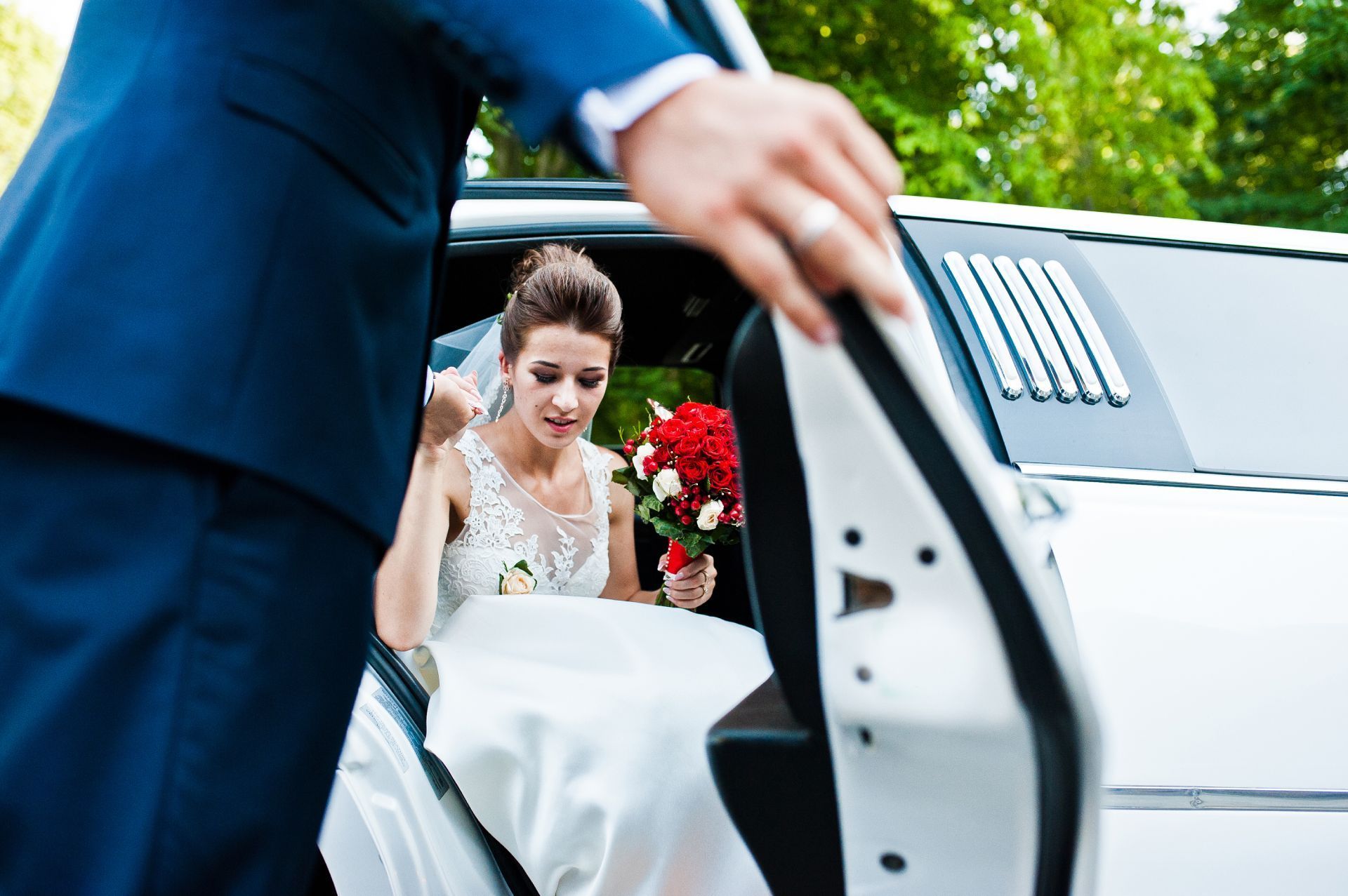 Bride exiting a white limousine; a man in a suit helps. She holds a bouquet.