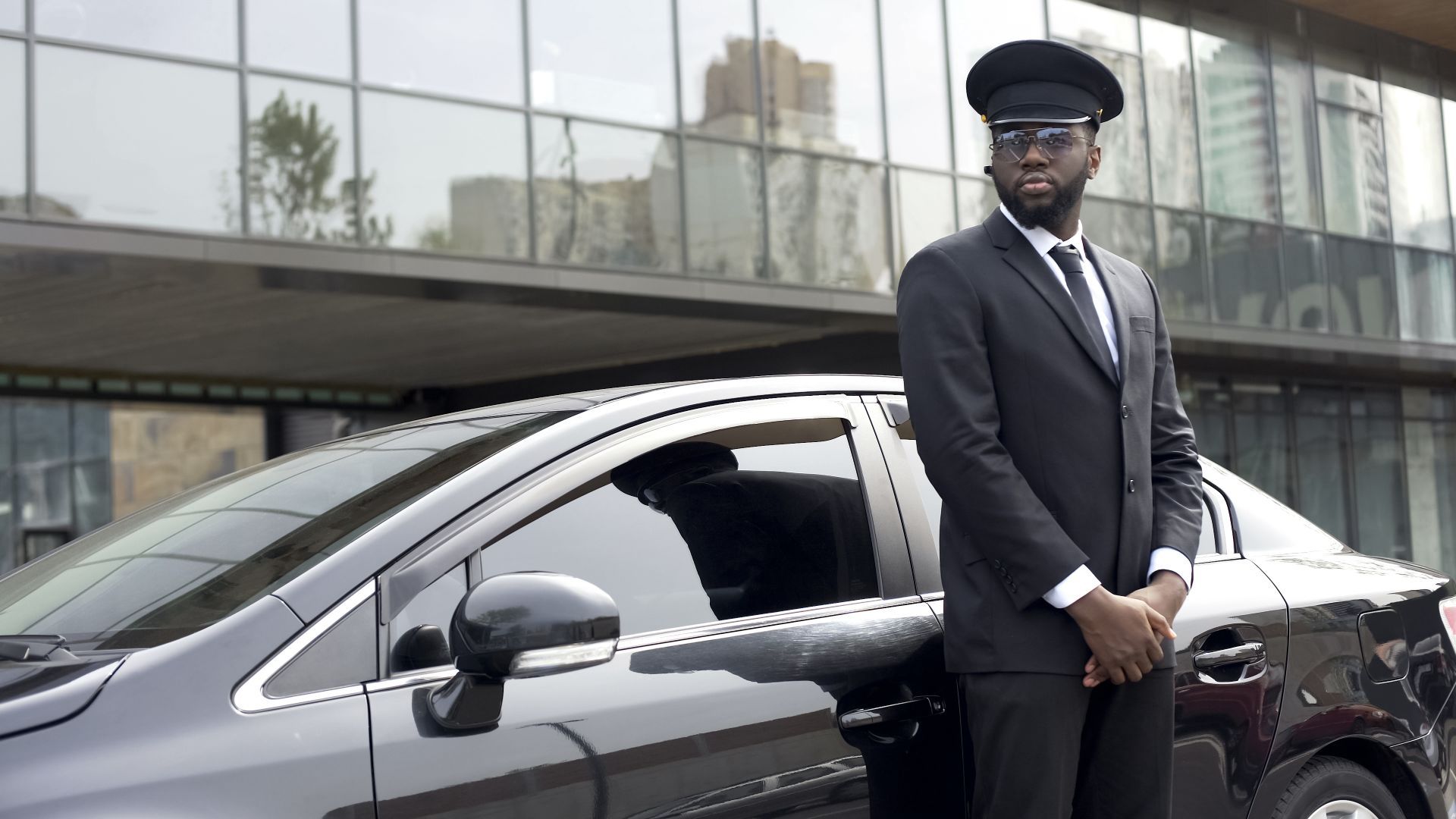 A Black chauffeur in a dark suit and hat stands next to a black sedan in front of a modern building.