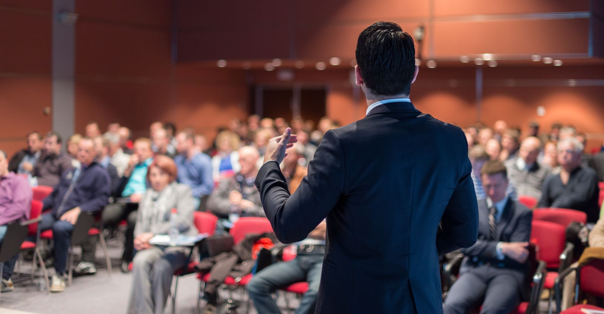 Man in suit speaking to a crowd at a conference, gesturing with his hands. Red chairs, wood paneling.