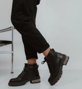 Persona con pantalones y botas negras. Un pie está inclinado hacia arriba. Las botas tienen suela gruesa.