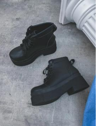 Botas de plataforma negras sobre una superficie gris cerca de un elemento arquitectónico blanco.