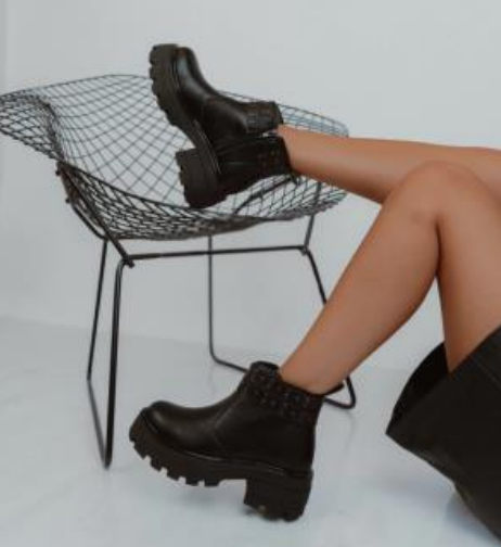 Mujer con chaqueta negra y botas de plataforma sentada en una silla, mirando fuera de cámara.