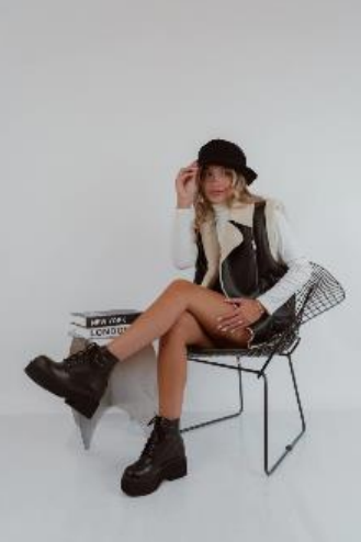 Mujer sentada en una silla, vestida con un chaleco negro, botas y sombrero, con un fondo neutro.