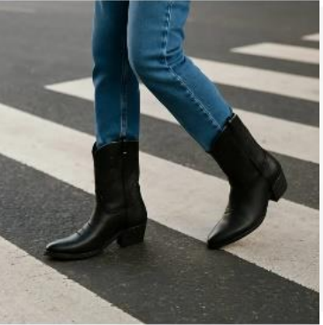 Persona con jeans azules y botas de vaquero negras caminando por un cruce de peatones.