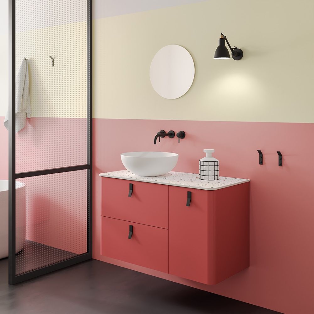 Bagno con mobiletto color corallo, pareti color crema, specchio rotondo e sanitari neri.