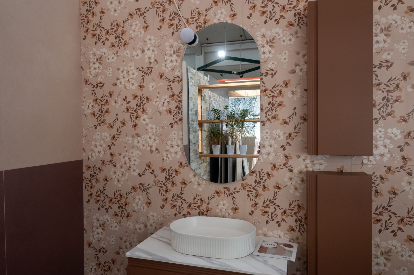 Bagno con specchio ovale, carta da parati floreale, mobili marroni e lavandino bianco.