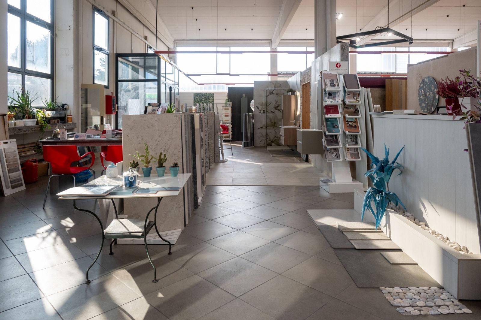 Vista interna di uno showroom con espositori di piastrelle, un tavolo con piante e ampie finestre.