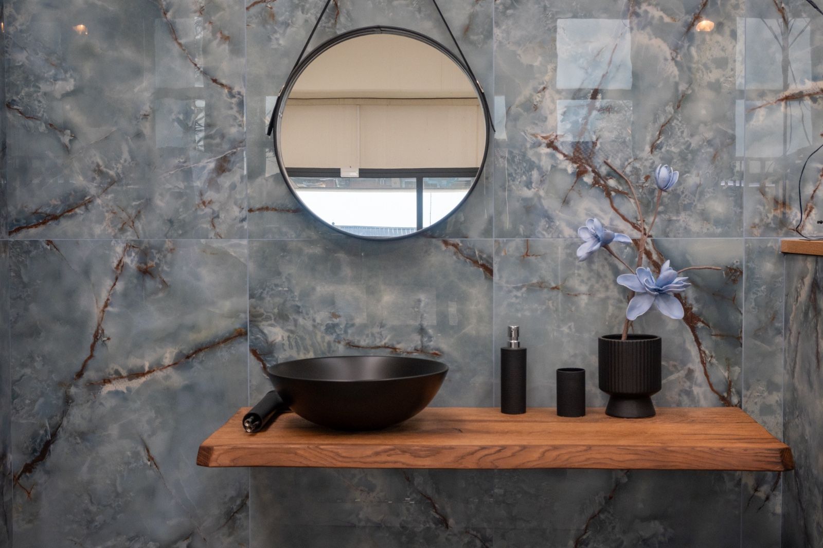 Bagno con parete piastrellata effetto marmo blu, lavandino e specchio neri, mensola in legno e oggetti decorativi.