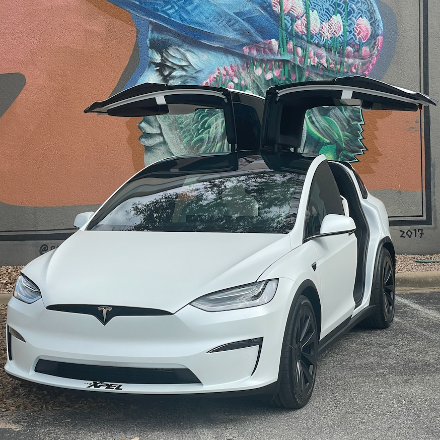 A white tesla model x with its doors open is parked in front of a mural.