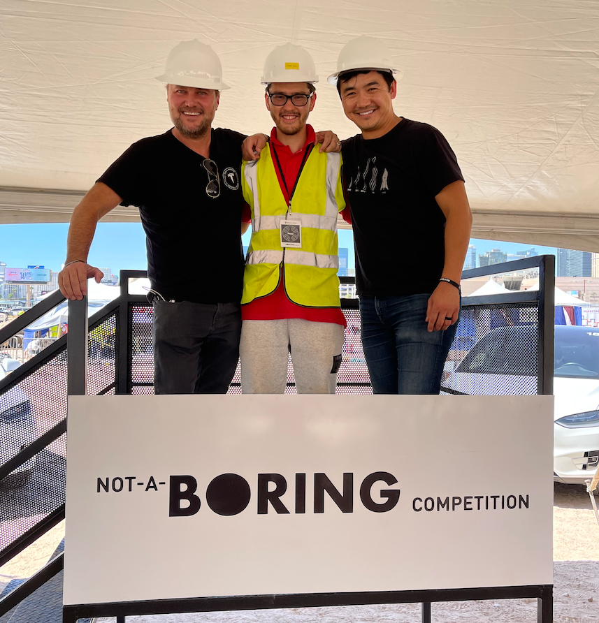 Three men standing next to a sign that says not a boring competition